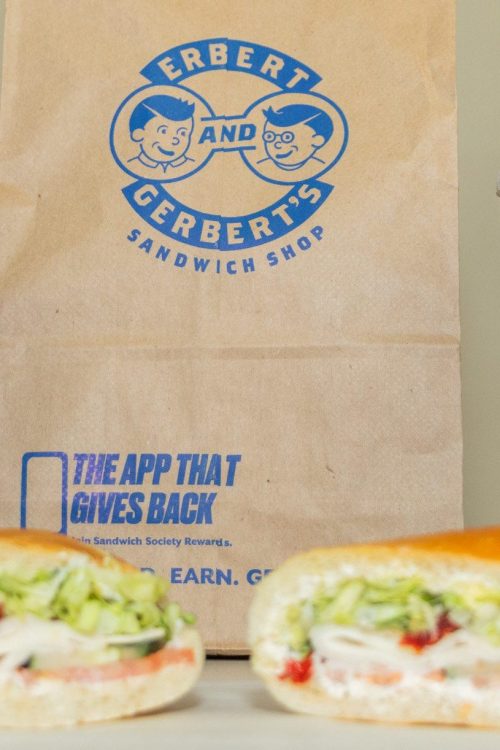 Two sandwiches cut in half with Pepsi drink cups in front of an Erbert and Gerbert bag.