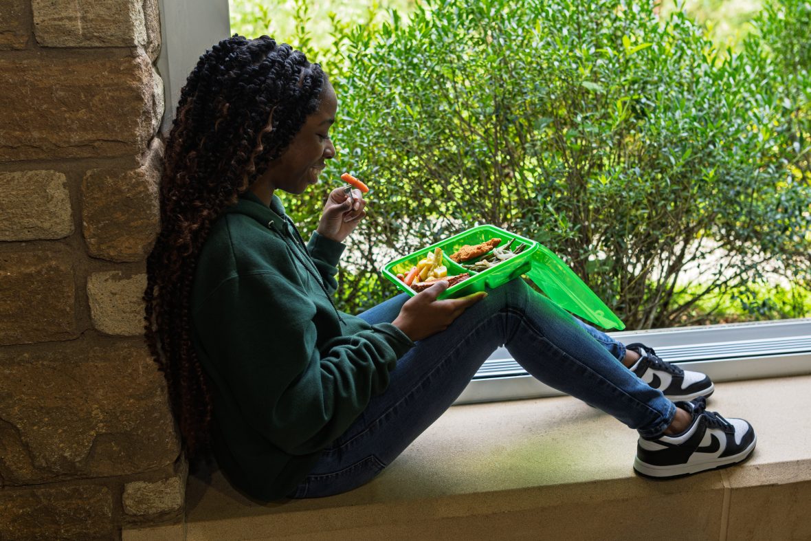 Student sitting on window ledge eating food out of green reusable container