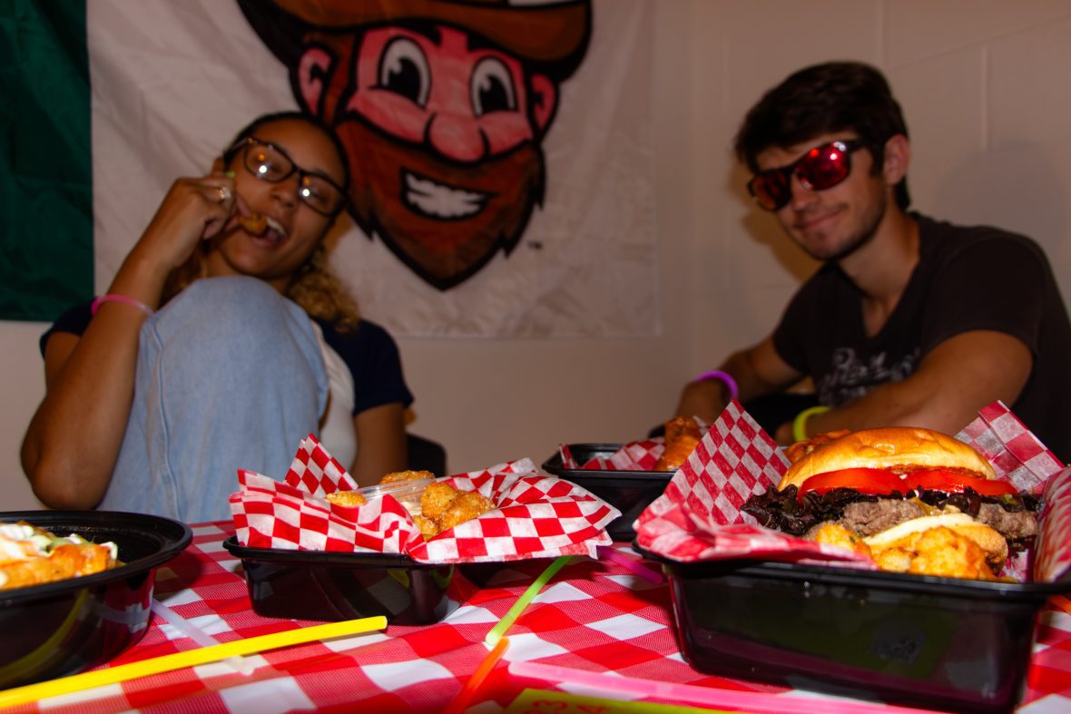 Baskets of fried food and burgers on table with glowsticks and two students wearing sunglasses in background