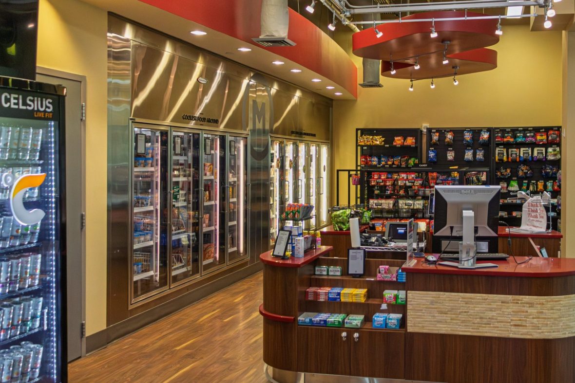 Inside of a marketplace with variety of drinks and snacks to grab, and check-out counter in middle of room