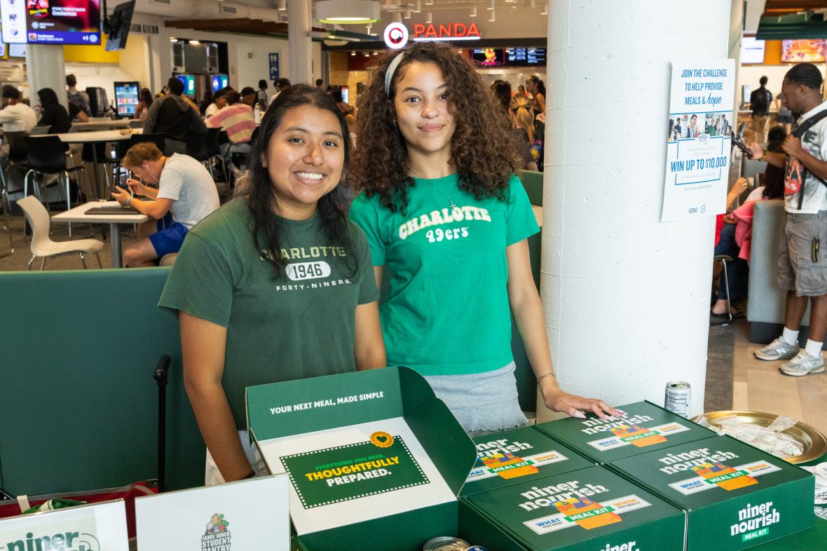 two students stand behind a table covered in Niner Nourish meal kit boxes